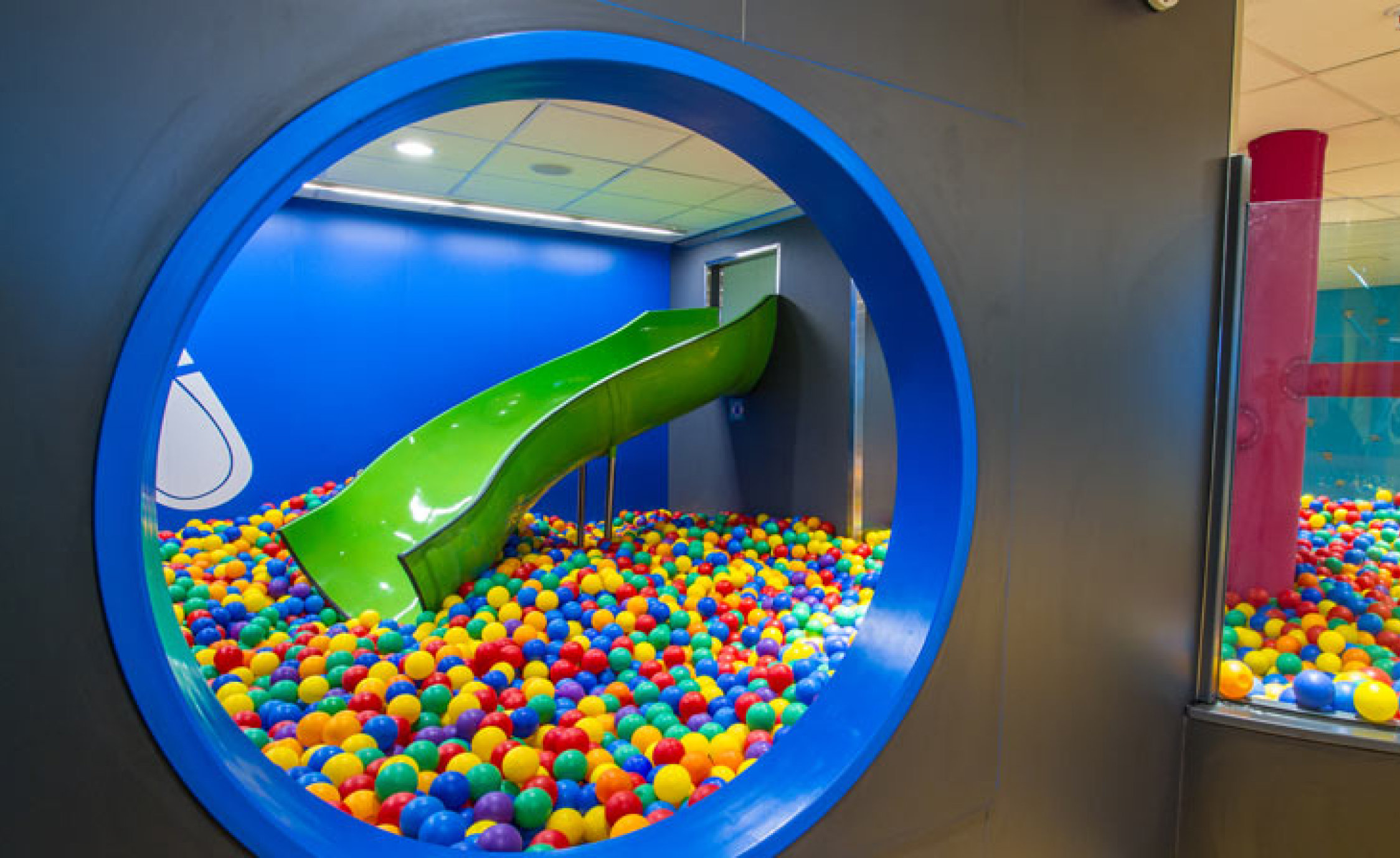En grön rutschkana som leder ner i ett färgglatt bollhav, sett genom ett runt fönster.