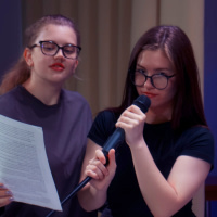 Två unga kvinnor med glasögon som sjunger karaoke och läser texter från ett papper.