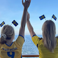 Två personer med gulblå fotbollströjor och händerna i luften tittar ut över havet