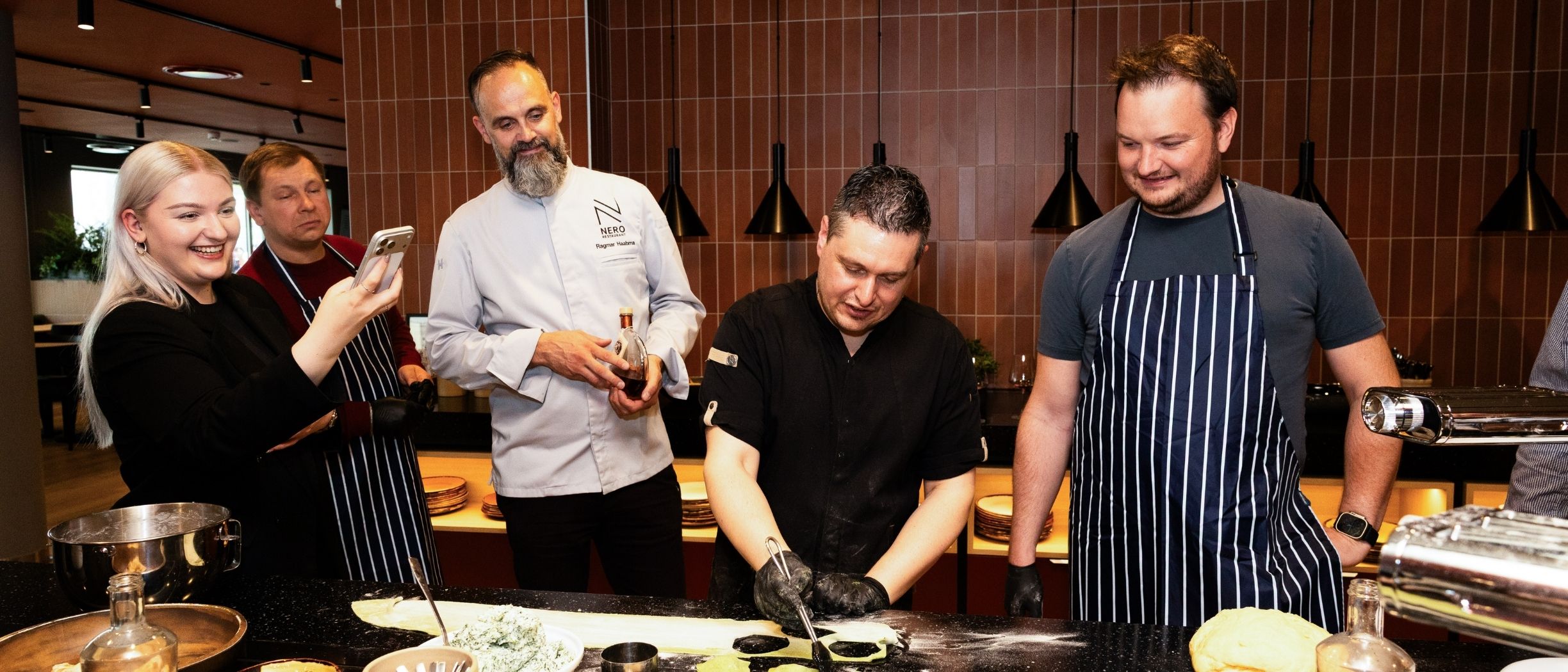 A group at a cooking class. Under the guidance of two chefs, pasta is being prepared while one guest takes photos of the experience.