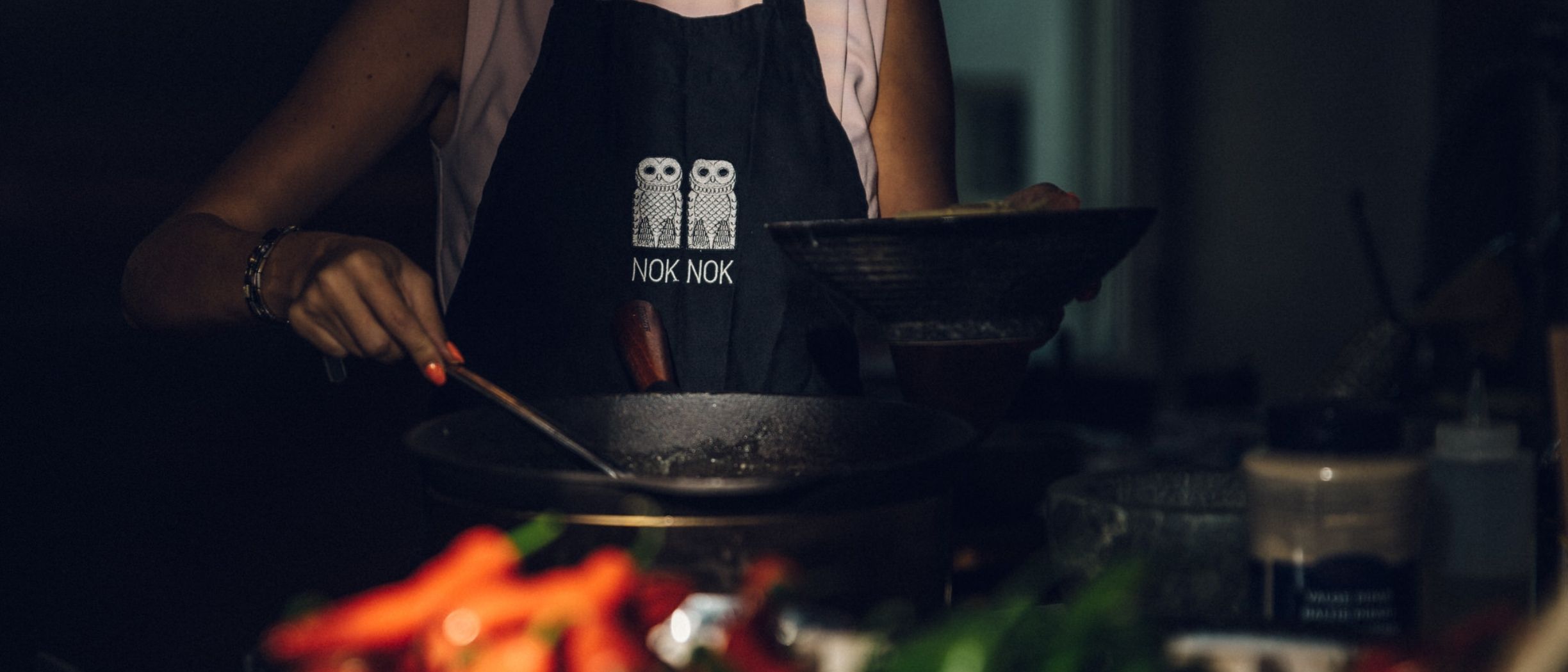 A guest at NOK NOK’s cooking school preparing food behind a wok pan, with red and green chilies vaguely visible in the steam.
