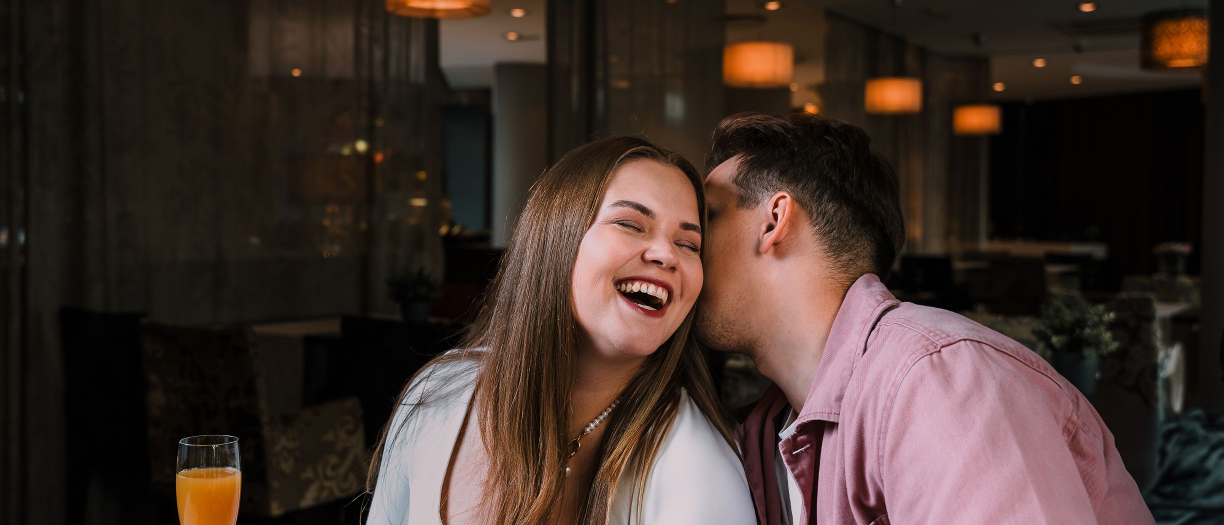 Mees ja naine puhkusel Riias, taustal on tumedates toonides hotelli sisemus. Naine naerab südamist, sest mees sositab talle miskit humoorikat kõrva sisse.