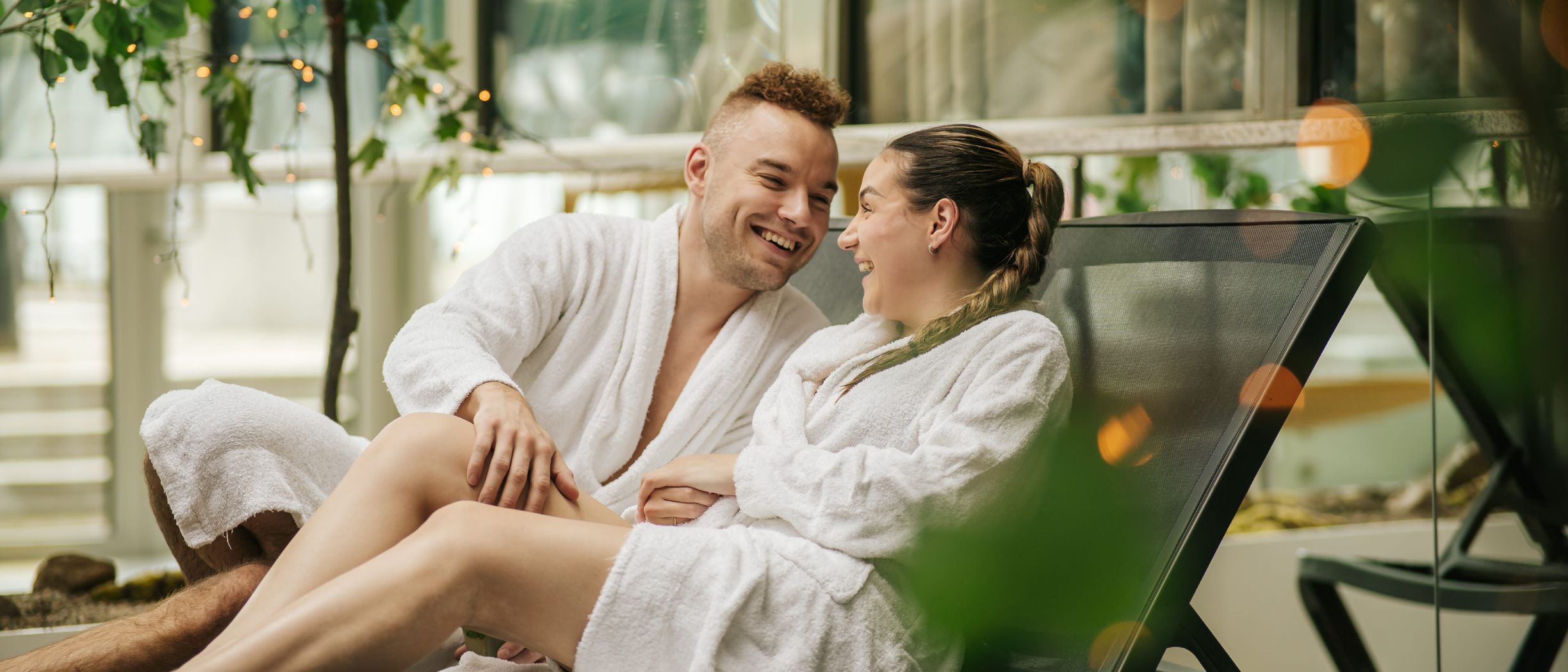 A young couple is enjoying each-others company. The are looking into each-others eyes and there are green plants in the background.