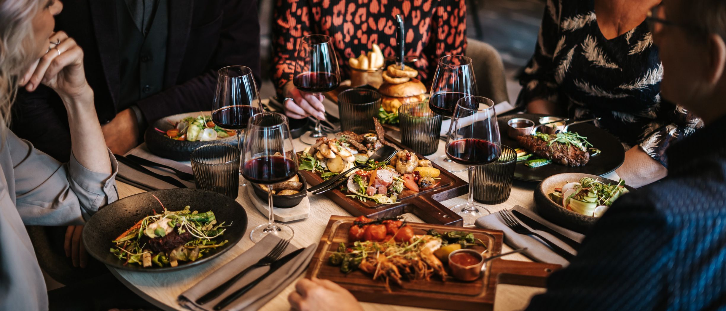 Dinner table with glasses of red wine and sharing dishes on wooden boards, with guests gathered around.