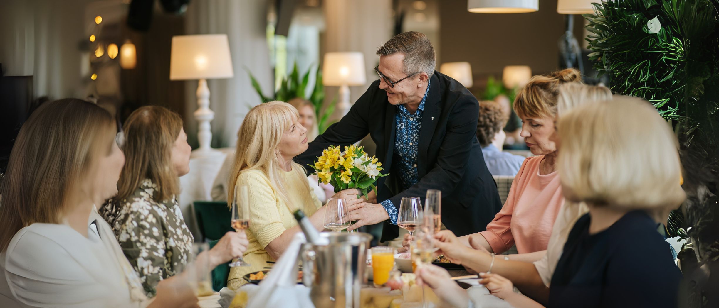 Six people sitting at a table, a dark haired man is giving a boquet of yellow flowers to a blonde woman