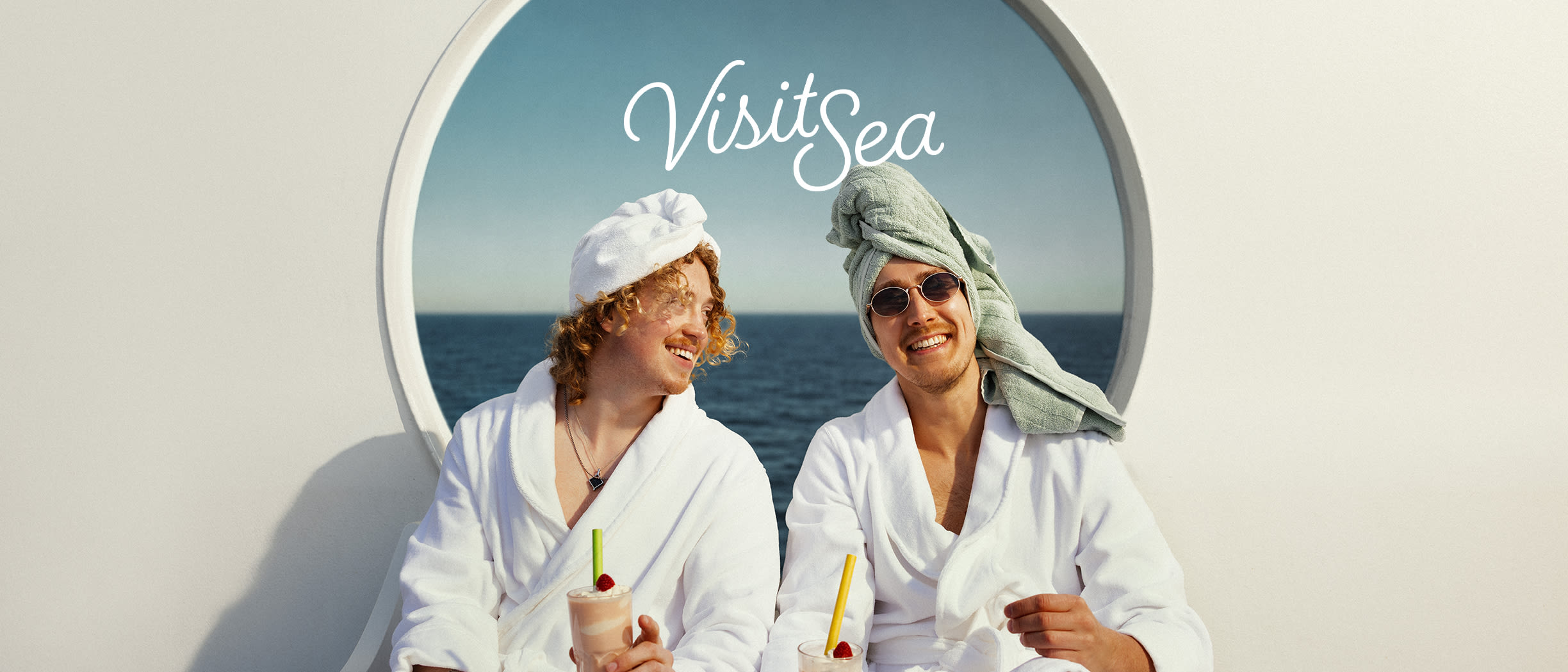 Two men in bathrobes and head towels relax with drinks on a ship. Text: VisitSea