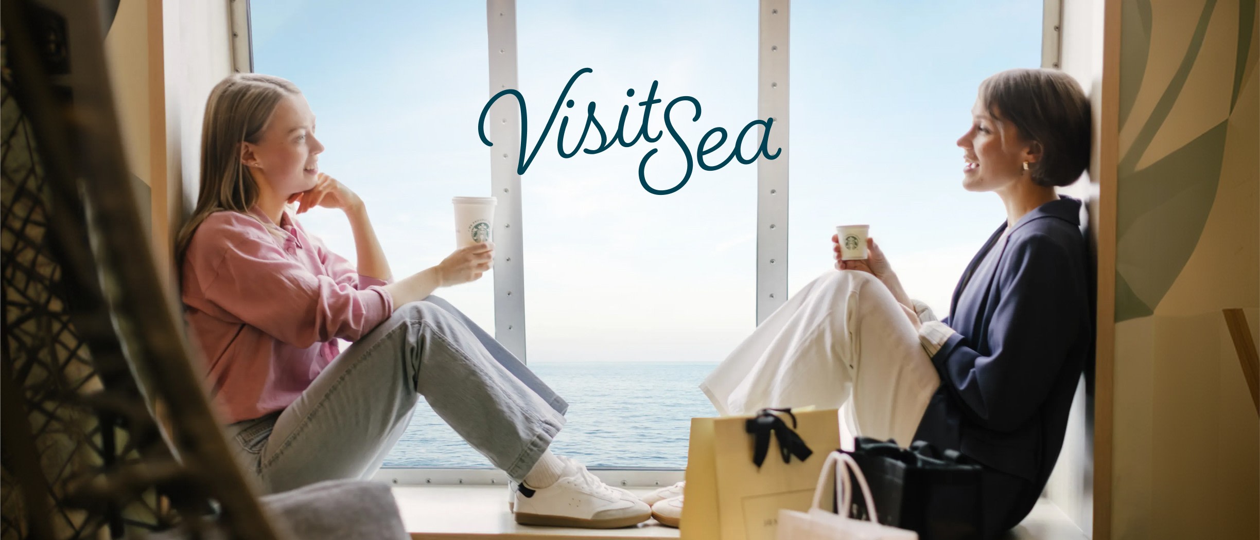 Divas sievietes sarunājas ar kafijas krūzēm pie loga ar skatu uz jūru, virs viņām logotips 'VisitSea'.
