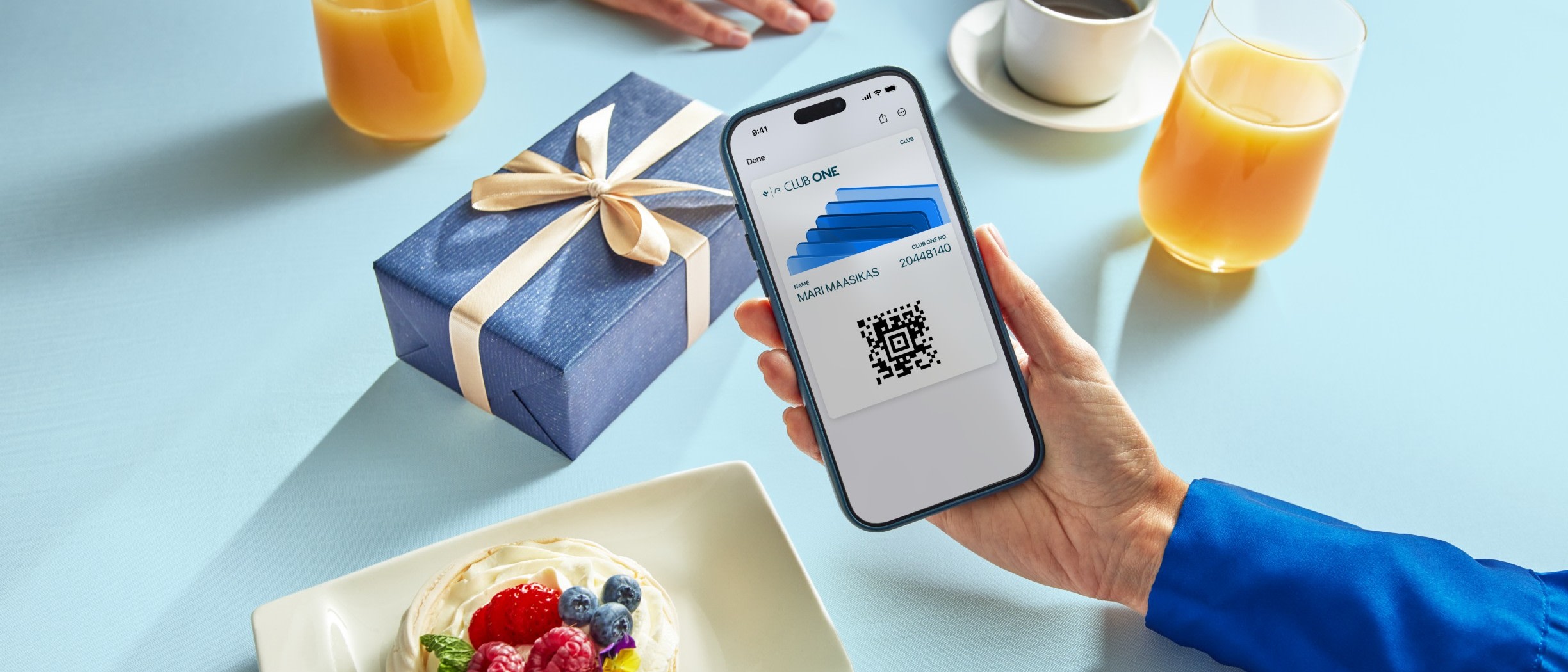 Hand holding a phone displaying a Club One digital loyalty card next to a gift and breakfast