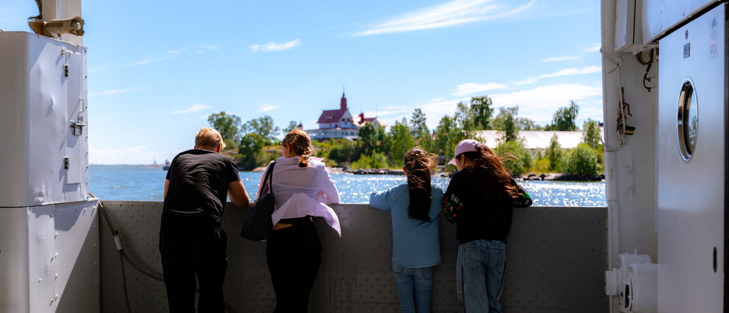 En familj står ute på däck på ett fartyg som åker förbi en liten ö i Helsingfors en solig sommardag