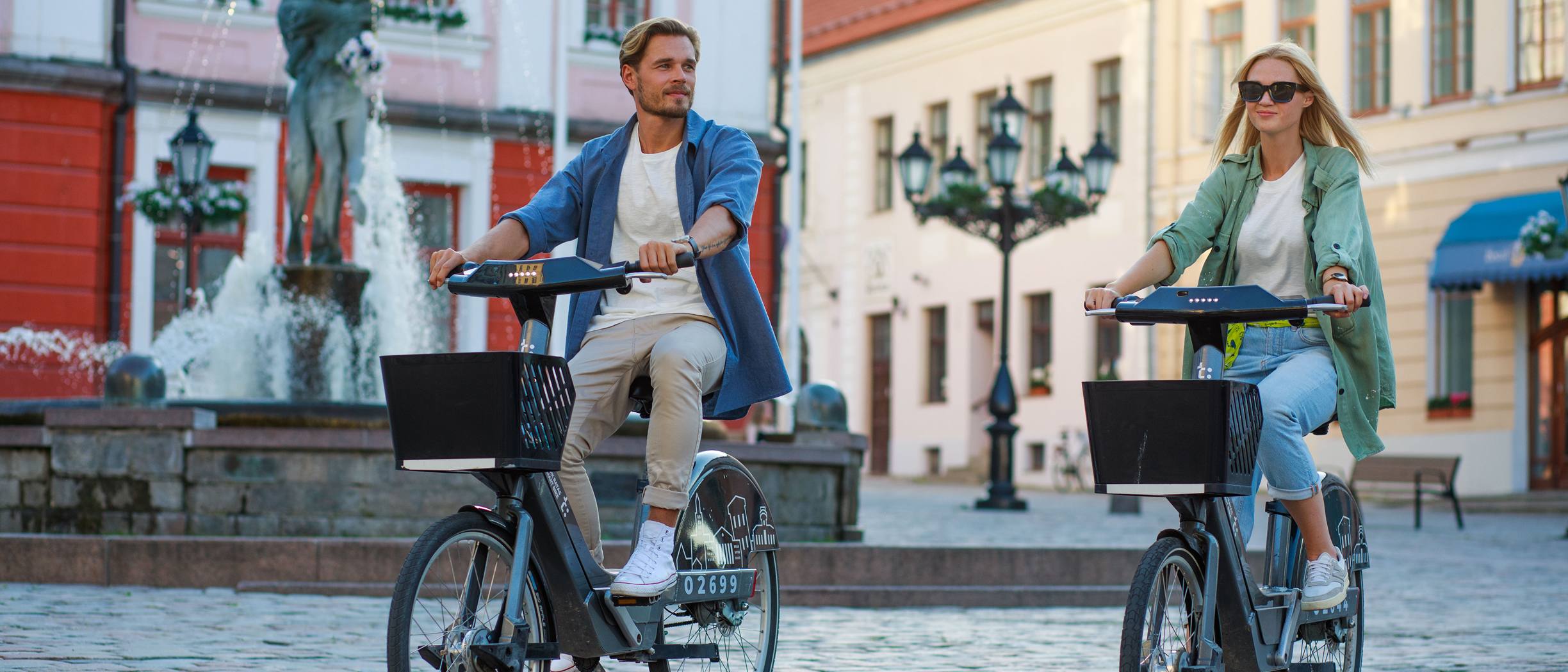 Couple cycling in Tartu Old Town near the Town Hall fountain on a sunny day.