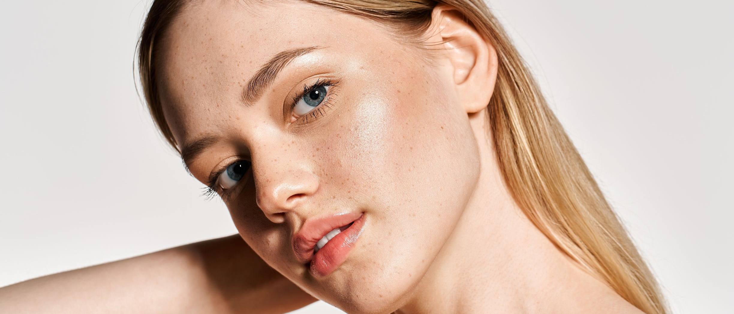 A young woman with fair skin and freckles is turned slightly to the side, looking at the camera. Her hair is sleekly pulled back and her skin has a natural, radiant glow.