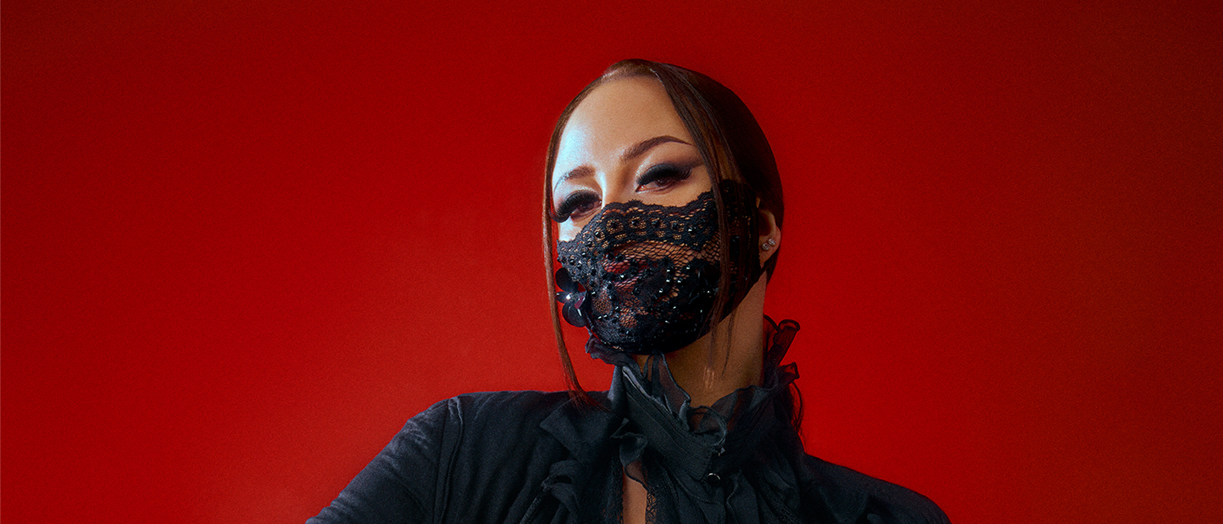 Felicia med dramatisk sminkning och svart spetsmask prydd med pärlor, mot röd bakgrund.
