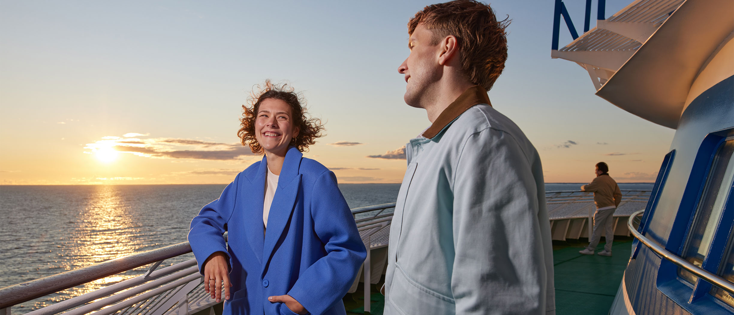 En tjej i blå kappa och en kille ute på däck och tittar på solnedgången