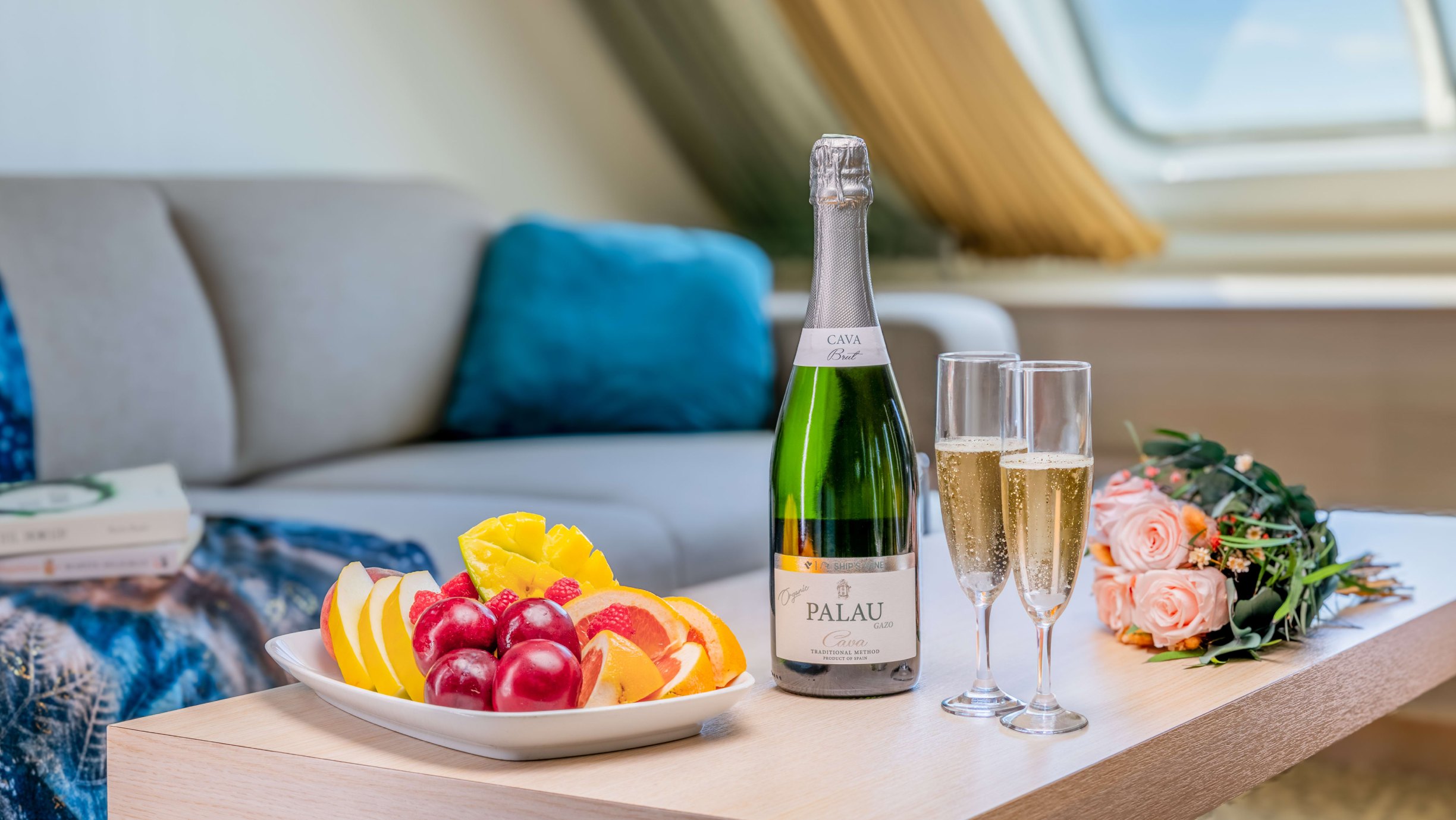Olika frukter, en flaska mousserande vin med två glas och en blombukett står på ett bord i en hytt