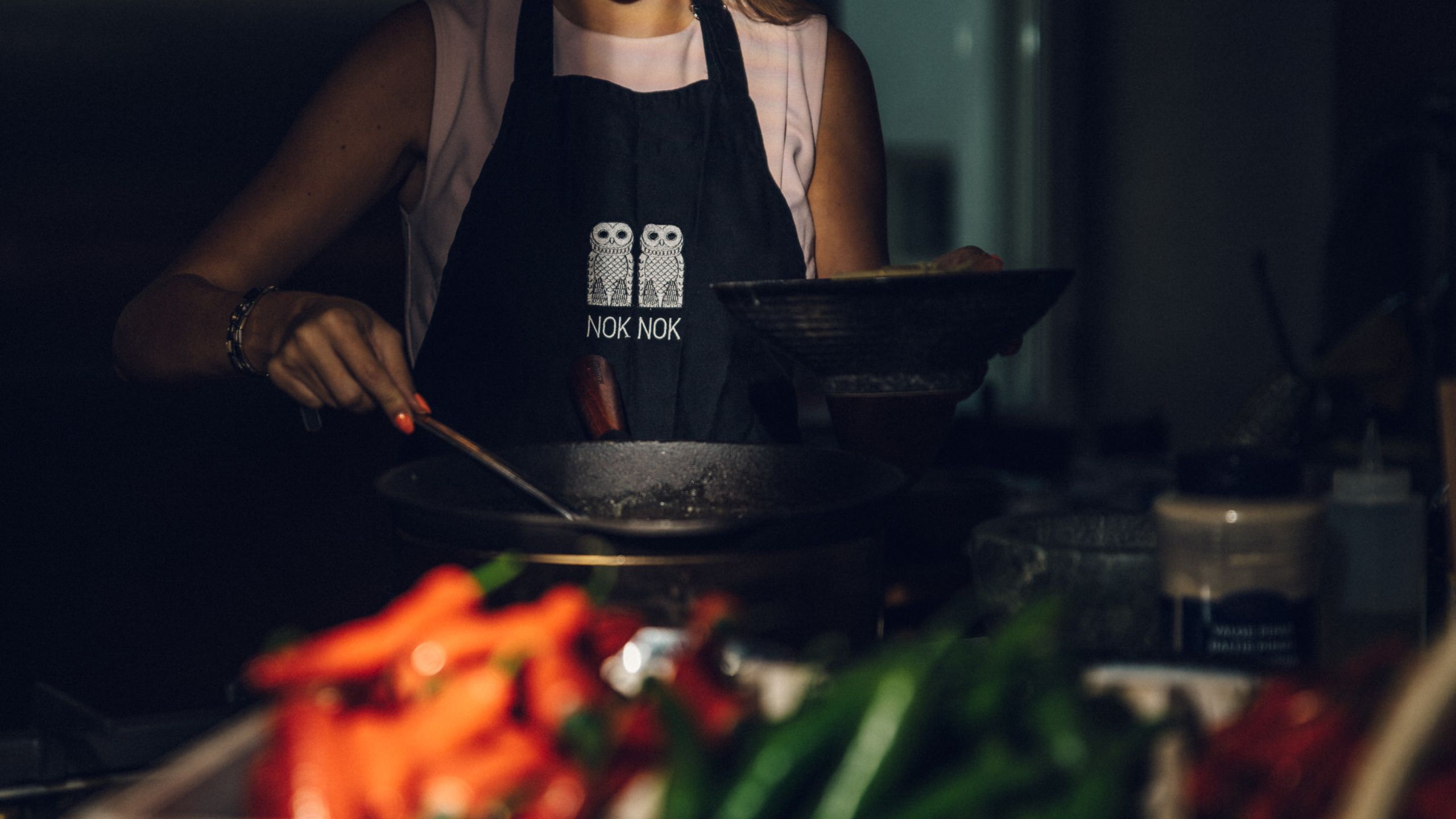 A guest at NOK NOK’s cooking school preparing food behind a wok pan, with red and green chilies vaguely visible in the steam.